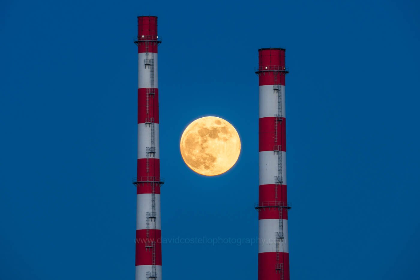 Full golden moon centred between the red and white Poolbeg Chimneys in Dublin Bay beneath a deep blue twilight sky.