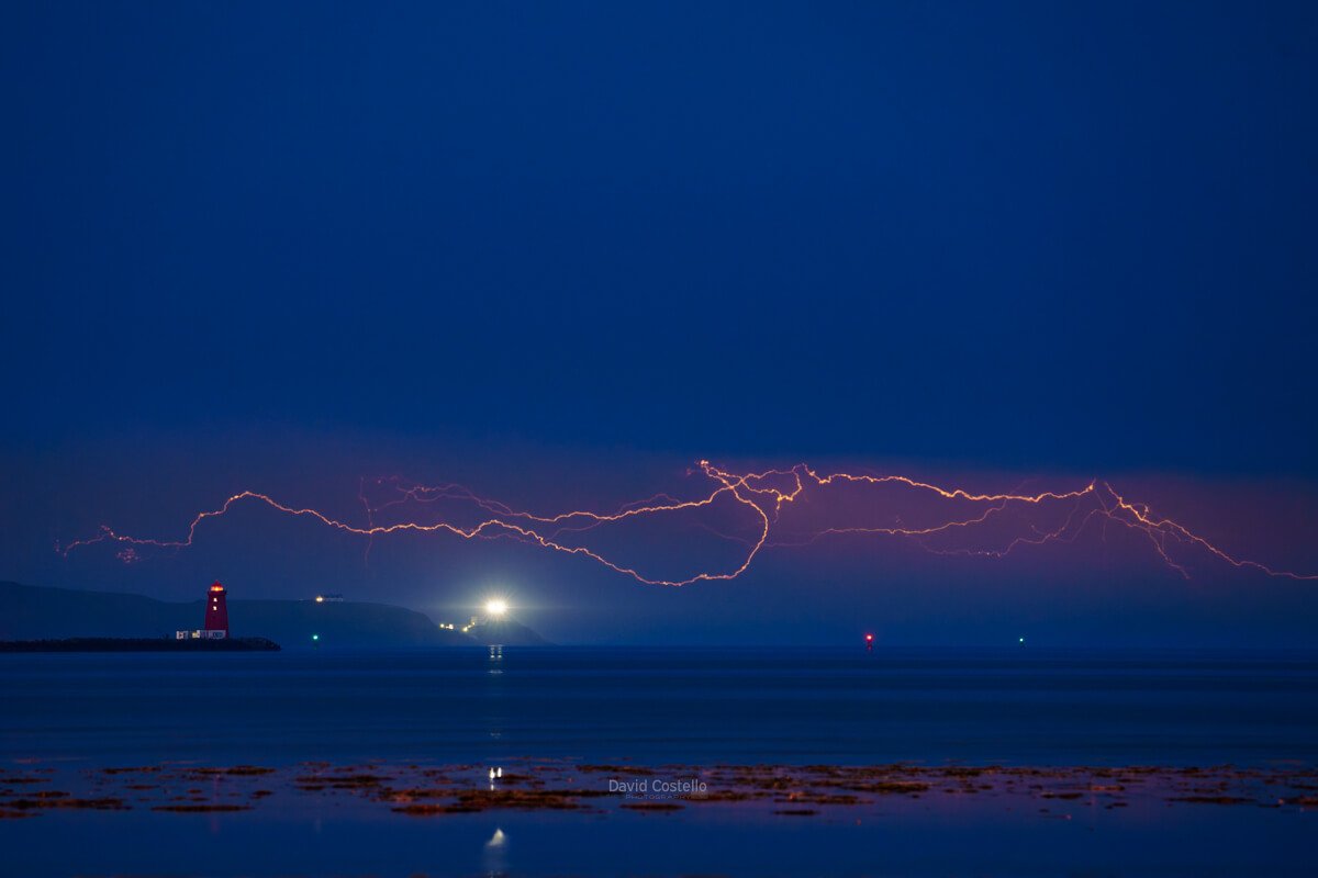 Lightning streaks across Dublin Bay above Poolbeg Lighthouse, with Baily Lighthouse visible on Howth Head, viewed from Sandymount Strand
