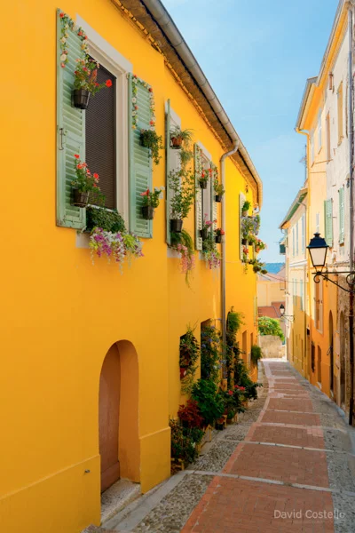 A bright yellow house with flowers in window boxes and hanging from the shutters along a Menton street.